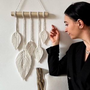 May include: A white macrame wall hanging with five leaf shapes, hanging from a wooden dowel rod. The wall hanging is made of cotton cord and is a popular bohemian decor item.