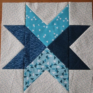 May include: A blue and white patchwork quilt block with a star design. The star is made up of four different blue fabrics with white accents. The background fabric is a light cream with a small white pattern.