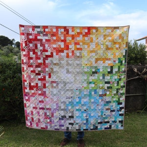 Pode incluir: Uma colcha de retalhos colorida com um padrão de arco-íris. A colcha é feita de quadrados de diferentes tecidos e cores, incluindo vermelho, laranja, amarelo, verde, azul, anil e violeta. A colcha está pendurada em um varal em um jardim.