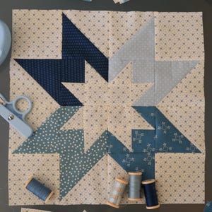 Pode incluir: Um bloco de colcha com um design de estrela em tons de azul e creme. O bloco é composto por quadrados e triângulos menores, e a estrela é formada pela disposição dos triângulos. O bloco é rodeado por um tecido creme com um pequeno padrão floral.