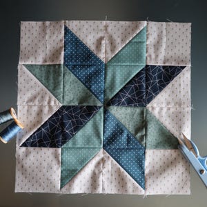 Può includere: Un blocco di trapunta patchwork con un motivo a stella in tonalità di blu, verde e bianco. Il blocco è realizzato con diversi tessuti con motivi diversi, tra cui pois, disegni geometrici e un tessuto bianco solido.