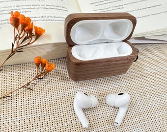 May include: A wooden case with a hinged lid, open to reveal two white compartments. The case is made of dark wood with a natural grain pattern. Two white wireless earbuds are lying on a woven surface next to the case.