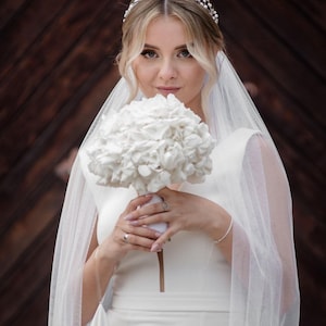 May include: A bride wearing a white wedding dress and veil holds a bouquet of white hydrangeas. She is wearing a silver tiara with pearls.