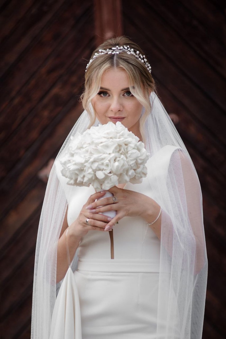 May include: A bride wearing a white wedding dress and veil holds a bouquet of white hydrangeas. She is wearing a silver tiara with pearls.