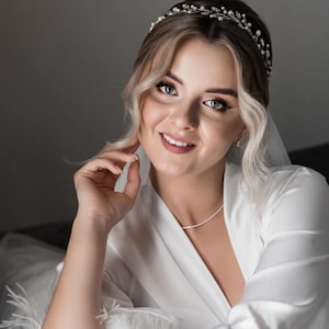 May include: A bride wearing a white satin robe with feather trim, a delicate silver headband with rhinestones, and a simple silver necklace. Her hair is styled in loose waves and she is smiling.