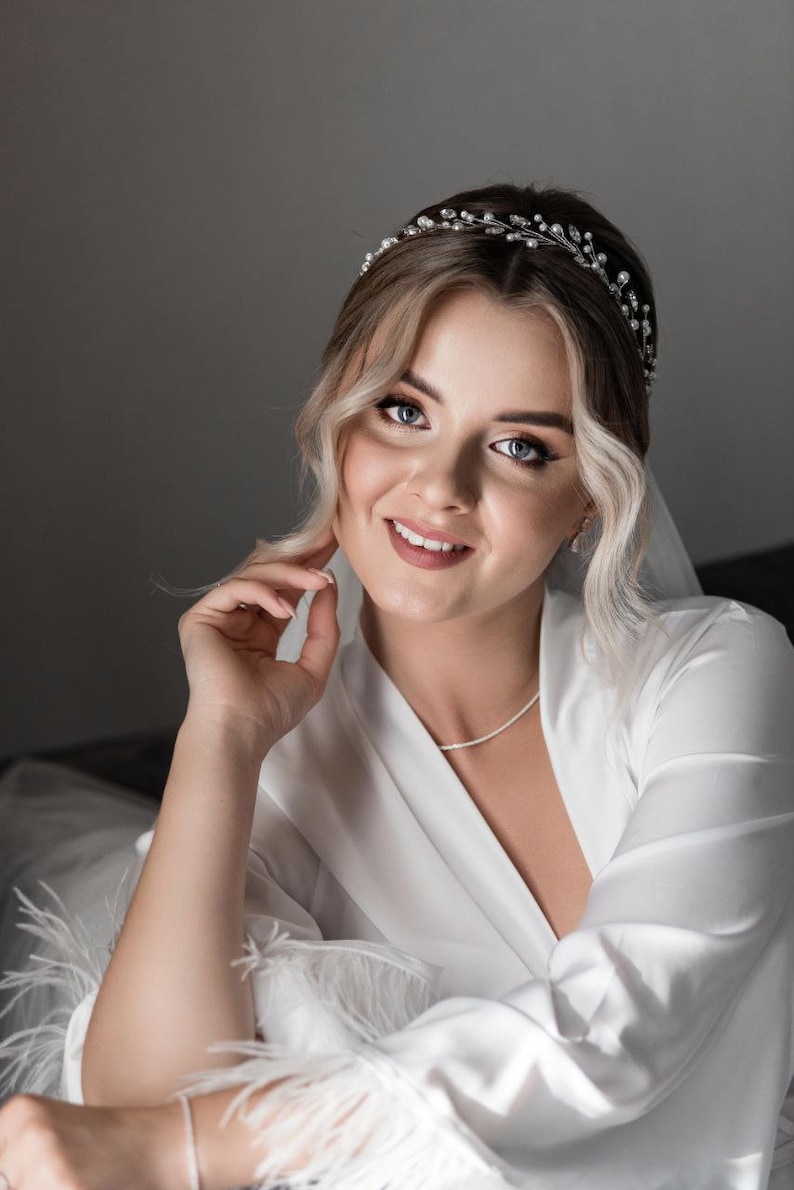 May include: A bride wearing a white satin robe with feather trim, a delicate silver headband with rhinestones, and a simple silver necklace. Her hair is styled in loose waves and she is smiling.