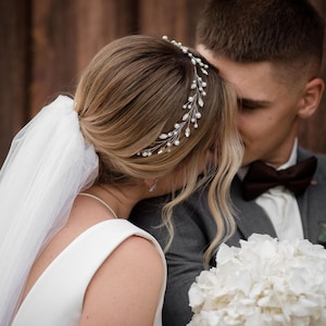 May include: A bride and groom are about to kiss. The bride is wearing a white dress and a veil. She has a silver tiara with pearls in her hair. The groom is wearing a gray suit with a brown bow tie. He is holding a bouquet of white flowers.