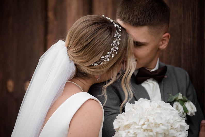 May include: A bride and groom are about to kiss. The bride is wearing a white dress and a veil. She has a silver tiara with pearls in her hair. The groom is wearing a gray suit with a brown bow tie. He is holding a bouquet of white flowers.