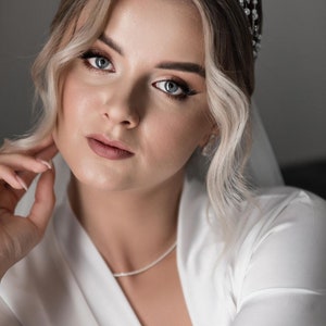 May include: A bride with blonde hair wearing a white robe and a pearl and silver headband. She is looking at the camera with a soft expression.