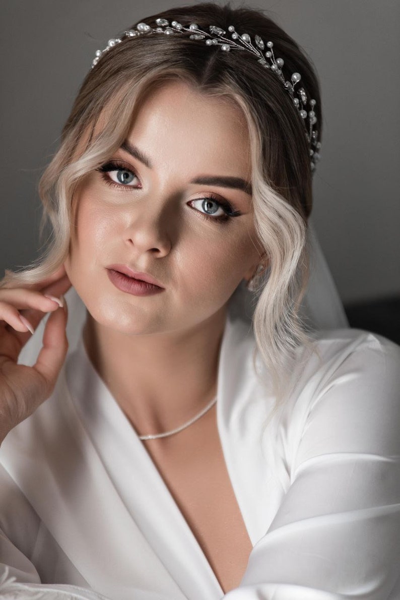 May include: A bride with blonde hair wearing a white robe and a pearl and silver headband. She is looking at the camera with a soft expression.