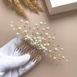 Könnte beinhalten: Ein goldfarbener Haarkamm mit Kunstperlen und Kristallen verziert. Der Kamm hat ein zartes Design mit Perlenclustern und funkelnden Akzenten, ideal für Braut- oder formelle Frisuren. Der Kamm wird von einer behandschuhten Hand gehalten.
