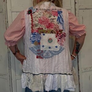 May include: A white and pink striped vest featuring a floral patchwork design on the back. The patchwork incorporates pink roses, blue and white patterned fabric, and lace trim. The vest has a ruffled hem and is worn over a pink blouse.