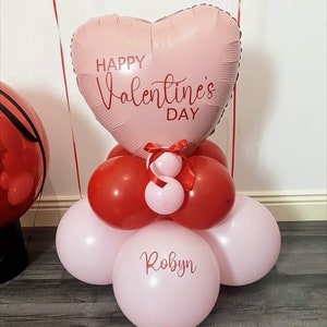 Puede incluir: Un globo rosa en forma de corazón con el texto "Happy Valentine's Day" en él. El globo está rodeado de globos más pequeños de color rojo y rosa. El globo inferior es rosa y tiene el nombre "Robyn" escrito en él.