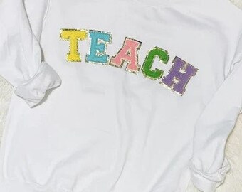 May include: White sweatshirt with the word "TEACH" in colorful, glittery letters.