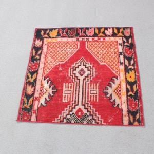 May include: A small rectangular rug featuring a vibrant red central field with a geometric mihrab design in cream, black, and orange. The border displays a repeating floral pattern in black, red, yellow, and pink against a dark background.