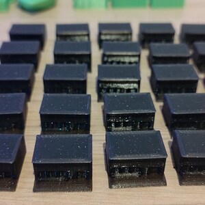 May include: A set of 24 black plastic miniature buildings, arranged in a grid pattern. The buildings are all the same size and shape, with a flat roof and a simple rectangular design.