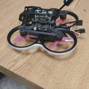 Puede incluir: Un dron negro con hélices rosas y un marco blanco. El dron está sentado en una mesa de madera.