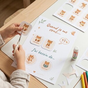 May include: A child is cutting out fox-themed emotion cards from a printed sheet. The sheet includes the phrases "Quand je me sens..." and "J'ai besoin de..." in French. Colored markers and glue are also on the table.