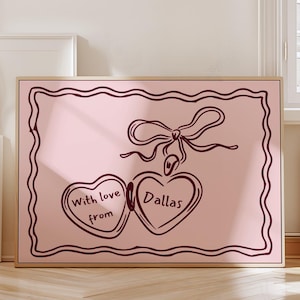Puede incluir: Impresión rosa enmarcada con dos corazones conectados, con la inscripción "With love from Dallas". La impresión tiene un borde marrón oscuro y un diseño de lazo en la parte superior.