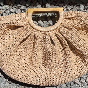 May include: A beige, woven handbag with a wooden handle. The bag has a rounded shape with a pleated design near the handle. A small, rectangular tag is attached to the side of the bag.