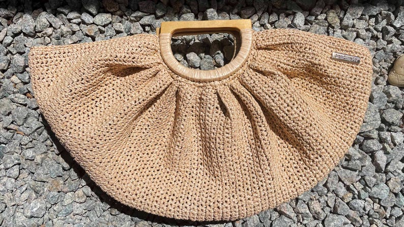 May include: A beige, woven handbag with a wooden handle. The bag has a rounded shape with a pleated design near the handle. A small, rectangular tag is attached to the side of the bag.