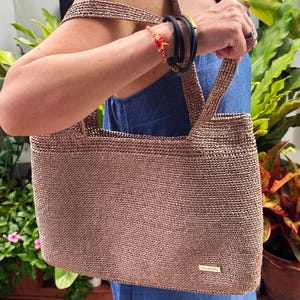 May include: A brown crocheted tote bag with two handles. The bag has a rectangular shape and a woven texture, with a small rectangular tag on the front. The bag is held by a person wearing a blue dress.