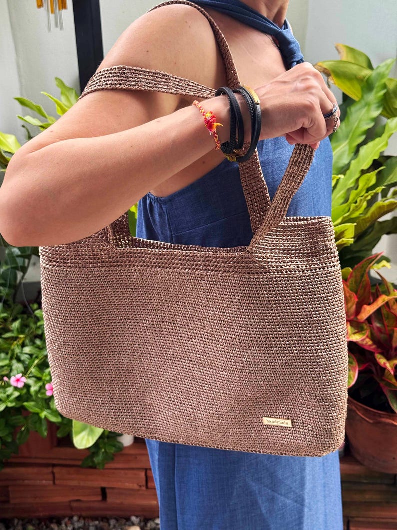 May include: A brown crocheted tote bag with two handles. The bag has a rectangular shape and a woven texture, with a small rectangular tag on the front. The bag is held by a person wearing a blue dress.