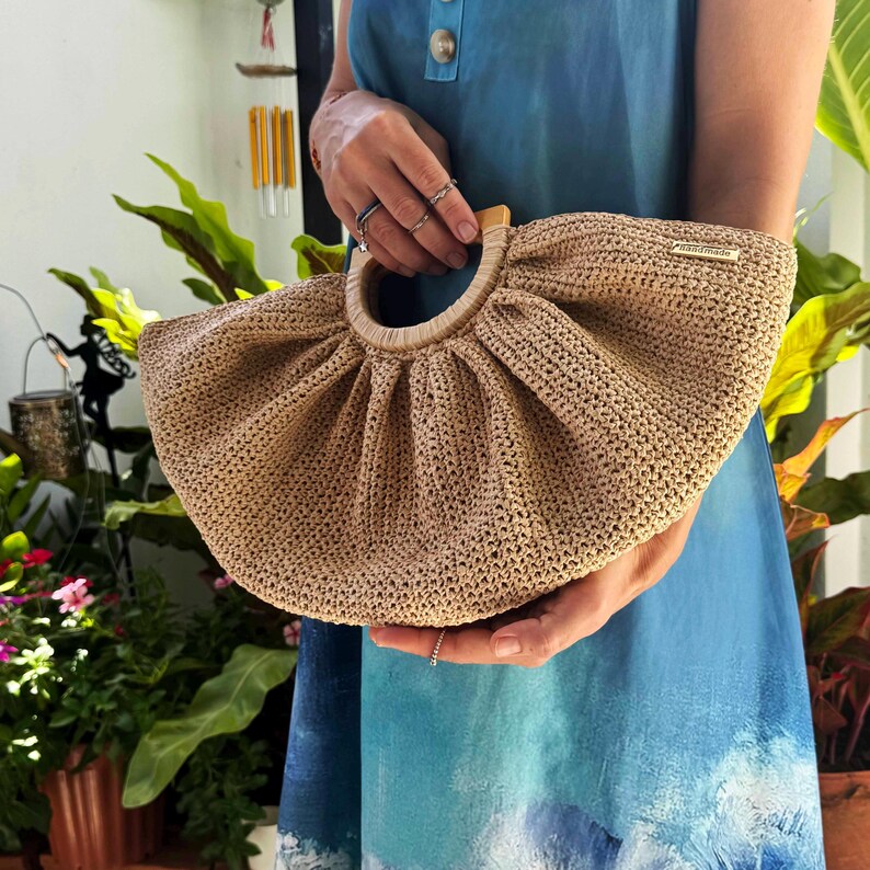 May include: A beige, hand-crocheted handbag with a wooden handle. The bag has a rounded shape and a gathered design. A small, gold-colored tag is attached to the top. The bag is held by a person wearing a blue dress.