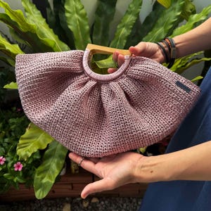 K&ouml;nnte beinhalten: Eine handgefertigte, geh&auml;kelte Handtasche in Staubrosa mit einem Holzgriff. Die Tasche hat eine runde Form und ein gerafftes Design an der Oberseite. Der Griff ist aus hellem Holz.