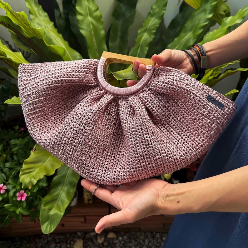 K&ouml;nnte beinhalten: Eine handgefertigte, geh&auml;kelte Handtasche in Staubrosa mit einem Holzgriff. Die Tasche hat eine runde Form und ein gerafftes Design an der Oberseite. Der Griff ist aus hellem Holz.