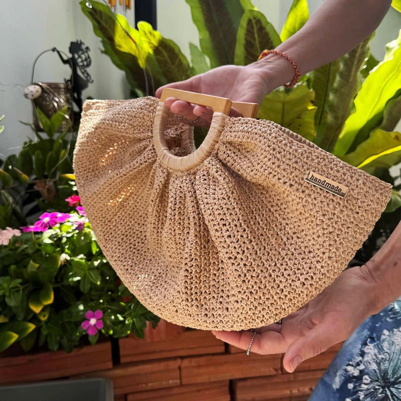 May include: A beige, handmade crocheted bag with wooden handles. The bag has a rounded shape and a label that reads "handmade". The bag is held in front of a background of green foliage and pink flowers.