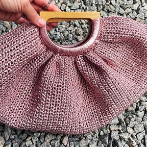 K&ouml;nnte beinhalten: Eine rosa Handtasche mit offenem Gewebe und einem Holzgriff. Die Tasche hat einen kreisf&ouml;rmigen Griff und ein gerafftes Design. Die Tasche besteht aus einem gl&auml;nzenden, strukturierten Material. Ein kleines schwarzes Etikett ist an der Seite der Tasche befestigt.