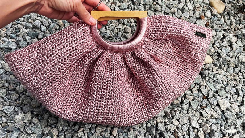 K&ouml;nnte beinhalten: Eine rosa Handtasche mit offenem Gewebe und einem Holzgriff. Die Tasche hat einen kreisf&ouml;rmigen Griff und ein gerafftes Design. Die Tasche besteht aus einem gl&auml;nzenden, strukturierten Material. Ein kleines schwarzes Etikett ist an der Seite der Tasche befestigt.