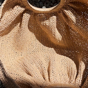 May include: A beige, open-weave tote bag with a circular wooden handle. The bag is made of a woven material, and the open weave creates a textured appearance. The bag is sitting on a surface of small, gray stones.