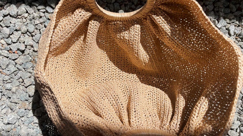 May include: A beige, open-weave tote bag with a circular wooden handle. The bag is made of a woven material, and the open weave creates a textured appearance. The bag is sitting on a surface of small, gray stones.