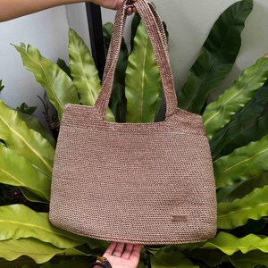 May include: A light brown woven tote bag with two long handles, held up by a person's hand. The bag has a rectangular shape and is made of a textured material. A small rectangular tag is visible on the front.