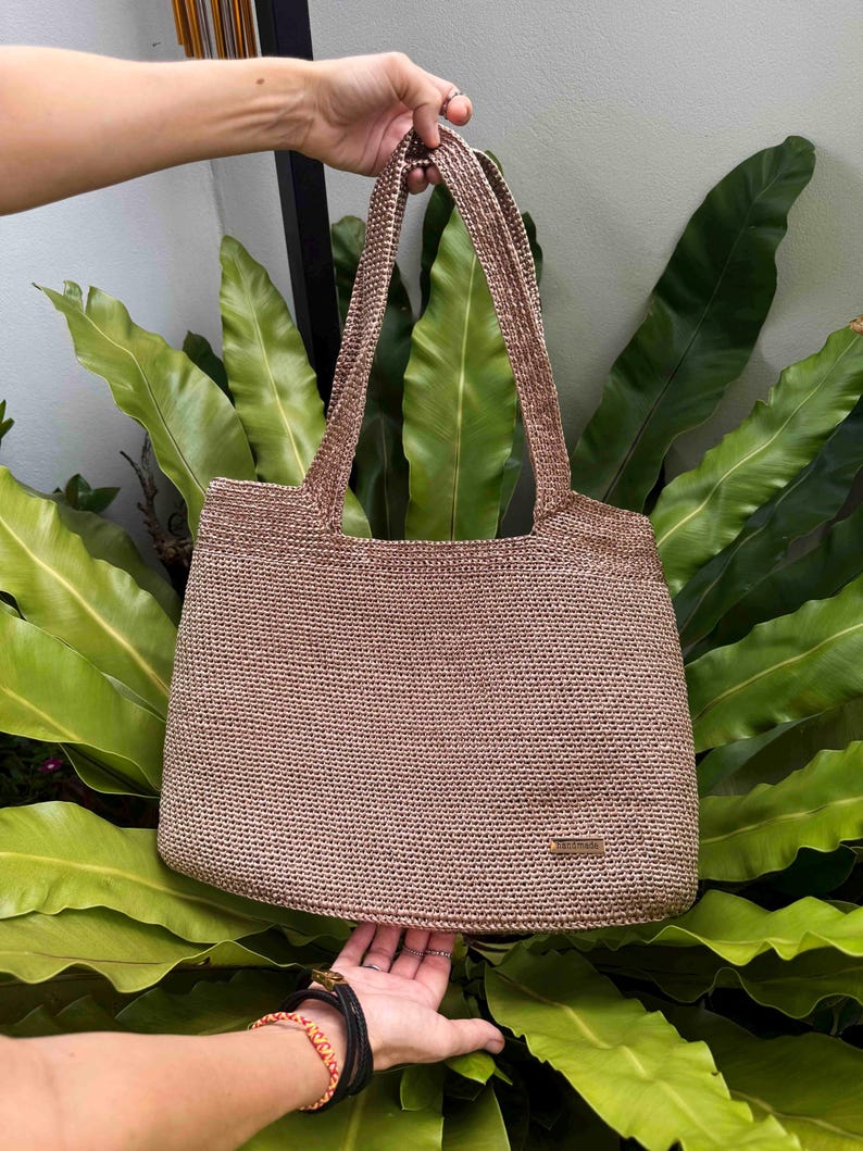 May include: A light brown woven tote bag with two long handles, held up by a person's hand. The bag has a rectangular shape and is made of a textured material. A small rectangular tag is visible on the front.