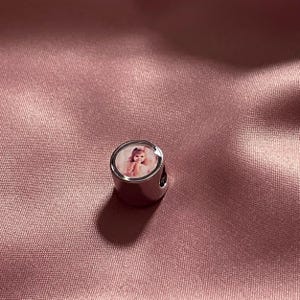Puede incluir: Un abalorio cil&iacute;ndrico de metal plateado que presenta una fotograf&iacute;a circular incrustada de un ni&ntilde;o peque&ntilde;o con cabello claro. El abalorio tiene una superficie pulida y reflectante con un orificio central para insertar una cadena.