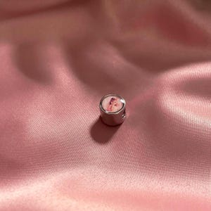 Puede incluir: Un charm cil&iacute;ndrico de metal plateado con una inserci&oacute;n circular en la superficie superior. La inserci&oacute;n muestra una peque&ntilde;a impresi&oacute;n fotogr&aacute;fica detallada de una persona de cabello claro, capturada en un estilo de retrato en tonos rosados.