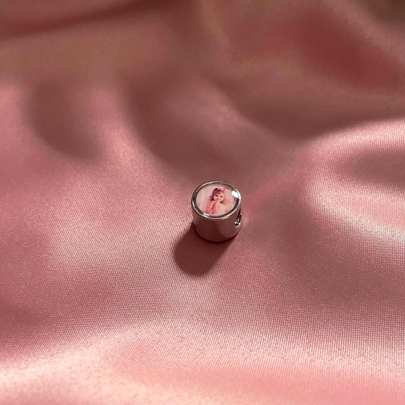 Puede incluir: Un charm cil&iacute;ndrico de metal plateado con una inserci&oacute;n circular en la superficie superior. La inserci&oacute;n muestra una peque&ntilde;a impresi&oacute;n fotogr&aacute;fica detallada de una persona de cabello claro, capturada en un estilo de retrato en tonos rosados.
