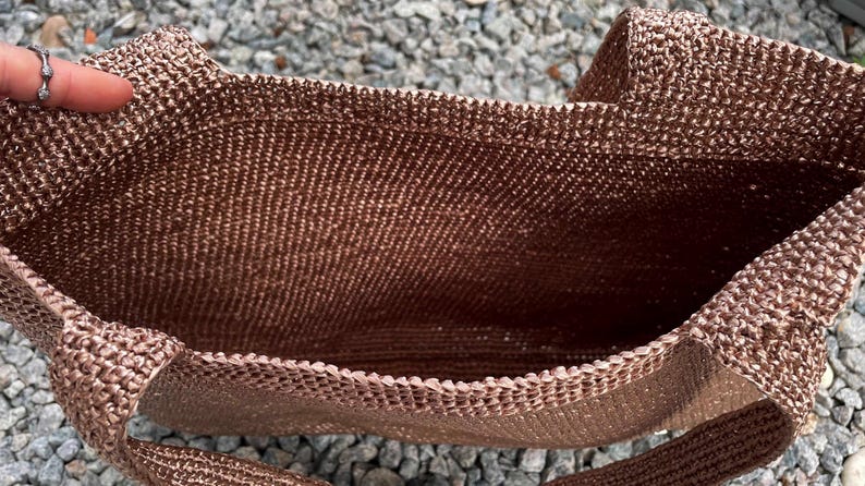 May include: A brown crocheted tote bag with a rectangular shape and two handles. The bag is made of a textured, woven material. The interior of the bag is visible, showing the open space for storage.