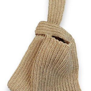May include: A gold-colored, handmade knitted bag with a knotted handle. The bag has a textured appearance and a small, rectangular tag that reads "handmade". The bag is designed with a unique shape and a spacious interior.