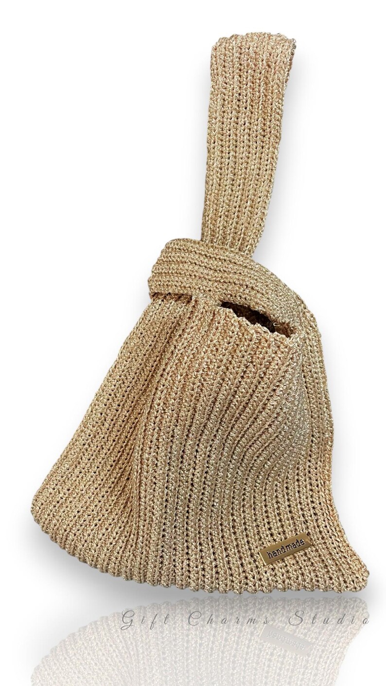 May include: A gold-colored, handmade knitted bag with a knotted handle. The bag has a textured appearance and a small, rectangular tag that reads "handmade". The bag is designed with a unique shape and a spacious interior.
