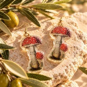 May include: A pair of dangle earrings featuring a clear resin design with a red and white mushroom illustration. Each earring has a gold-toned metal hook and displays a large mushroom with a smaller mushroom beside it, accented with subtle glitter.