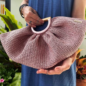 K&ouml;nnte beinhalten: Handgefertigte, altrosa geh&auml;kelte Handtasche mit Holzgriff. Die Tasche hat eine runde Form und ein zartes, offenes Webmuster. Der Griff ist aus hellem Holz, und die Tasche wird von einer Person mit Ringen und Armb&auml;ndern gehalten.