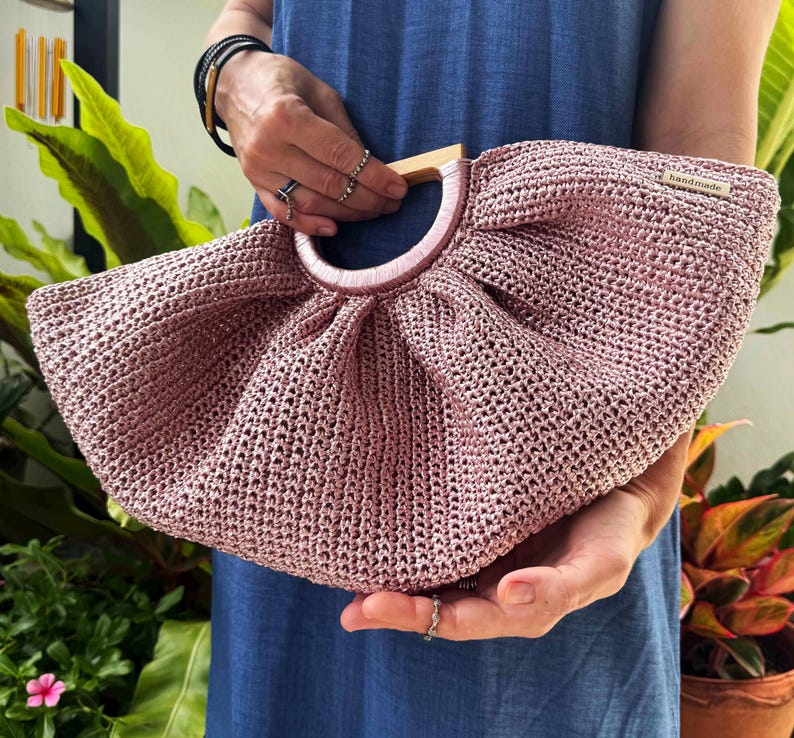 K&ouml;nnte beinhalten: Handgefertigte, altrosa geh&auml;kelte Handtasche mit Holzgriff. Die Tasche hat eine runde Form und ein zartes, offenes Webmuster. Der Griff ist aus hellem Holz, und die Tasche wird von einer Person mit Ringen und Armb&auml;ndern gehalten.