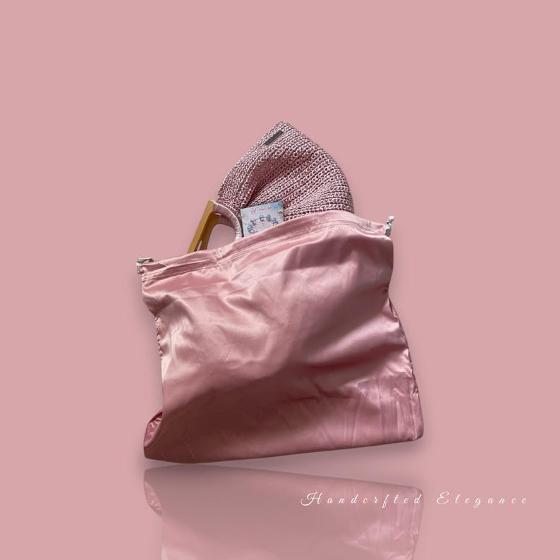 K&ouml;nnte beinhalten: Eine rosa Satin-Tragetasche mit einem gewebten rosa Hut im Inneren. Die Tasche hat einen Holzgriff und einen Kordelzugverschluss. Der Hintergrund ist einfarbig rosa. Die Worte "Handcrafted Elegance" sind unten in Kursivschrift geschrieben.