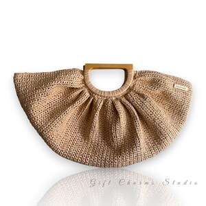 May include: A beige, handmade crochet handbag with a wooden handle. The bag has a fan-like shape and a small tag that reads "handmade". The bag is a neutral color and has a textured appearance.