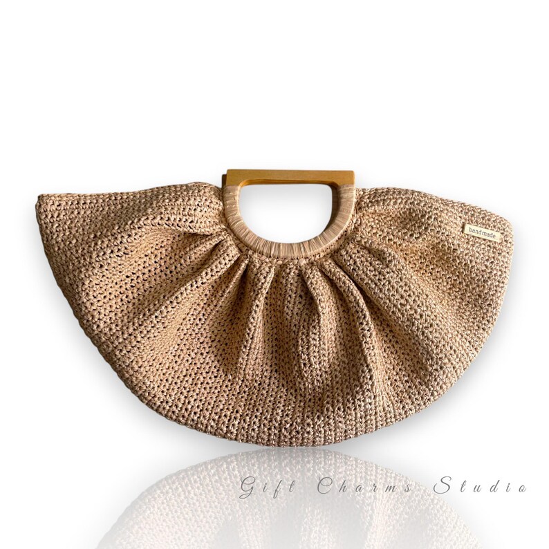 May include: A beige, handmade crochet handbag with a wooden handle. The bag has a fan-like shape and a small tag that reads "handmade". The bag is a neutral color and has a textured appearance.