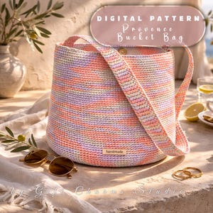 May include: A handmade, crochet bucket bag in pastel colors of pink, orange, and lavender. The bag has a matching strap and a small "handmade" tag. The image also includes sunglasses, gold rings, and text that reads "Digital Pattern Provence Bucket Bag."